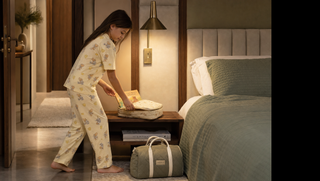 Girl in golden cream pyjama set placing a storybook into an overnight pouch in a warm guest room