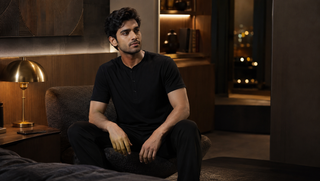 Synthetic Indian man in a black Sweet Dreams-style Henley pyjama set seated in a charcoal and walnut bedroom lounge, illustrating how sleep does not stop listening.