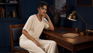 AI-generated woman in whipped cream wide-leg pyjama set seated at a walnut desk in a blue night study with warm brass lamp light