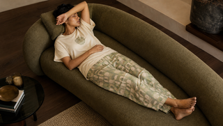 Synthetic South Asian woman in cream-and-sage sleepwear drifting across an olive sofa in late afternoon light, illustrating how a late nap can steal from bedtime.