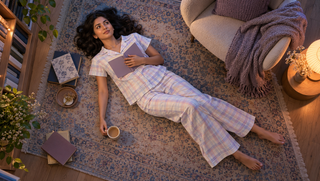 Synthetic Indian woman in pastel plaid palazzo pyjamas reflecting with a notebook on a library rug at dawn
