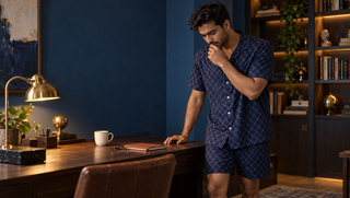 A synthetic man in navy geometric sleepwear pauses at a walnut study desk in the morning