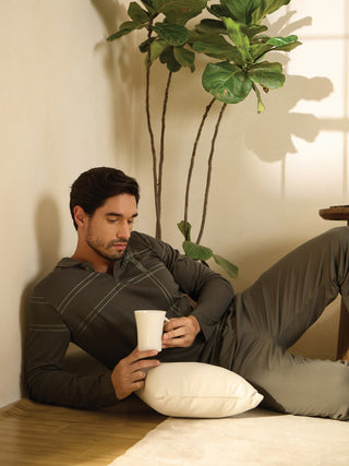 A man reclines on the floor, holding a mug and resting his head on a pillow, wearing a dark grey long-sleeve polo with a subtle plaid print and matching pants.