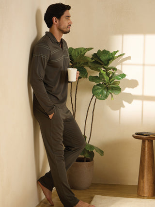 A man in a dark grey long-sleeve polo and matching pants leans against a wall, holding a white mug, a Fiddle Leaf Fig plant beside him.