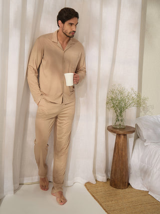 A man in beige pajamas stands holding a white mug next to a bedside table with a vase of flowers.