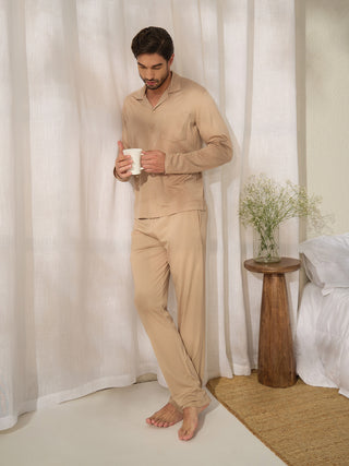 Men in a beige pajama set holding a mug by the window, with a small bouquet on a side table.
