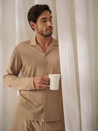 A man in a beige pajama set, holding a white mug, stands near a white curtain.