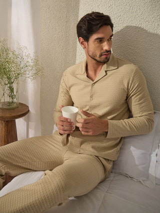 A man is sitting on a bed wearing a tan patterned pajama set and holding a white mug.
