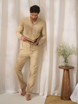 Men's beige textured pajama set, posed leaning against a curtain while reading, with a bouquet on a wooden table.
