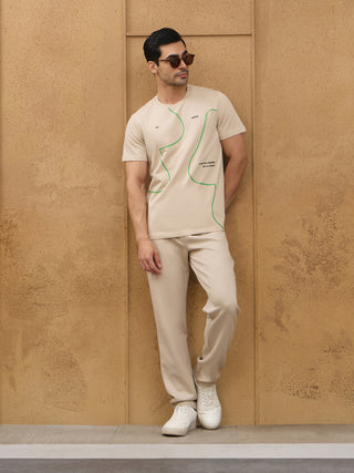 A man wears a beige t-shirt with green line art and matching beige pants, leaning against a wall.