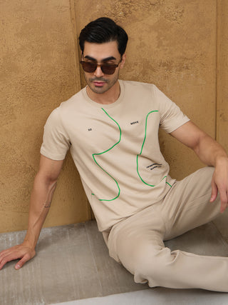 Men's light beige t-shirt with green abstract print and matching pants, posed leaning against a textured wall.