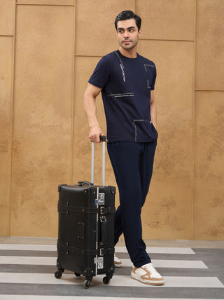 Men standing with a black suitcase, wearing a dark blue t-shirt with geometric print and dark blue pants.