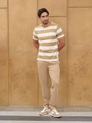 A man stands looking to the right, wearing a horizontal striped t-shirt in beige and white and beige trousers.
