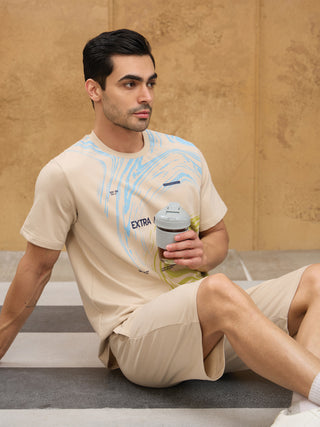 Men wearing a light beige t-shirt with a blue and green abstract print and matching shorts, sitting and holding a grey travel mug.