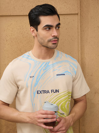 A man poses holding a coffee cup in a beige t-shirt with a blue and green abstract print.