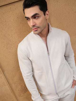 Men wearing a light cream full-zip loungewear jacket and pants, posing looking away from the camera against a textured brown wall.