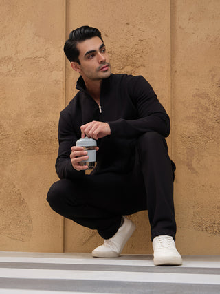 A man in a black zip-up jacket and pants squats holding a water bottle, with white sneakers.