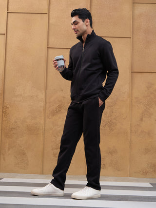 A man in a black zip-up jacket and pants stands holding a water bottle, with white sneakers.