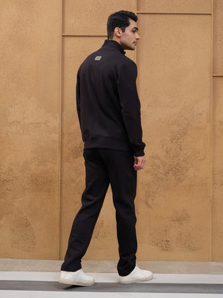 A man in a black zip-up jacket and pants stands, with white sneakers.