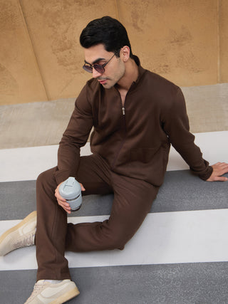 Men in a brown zippered sweatshirt and pants, sitting and holding a grey cup.