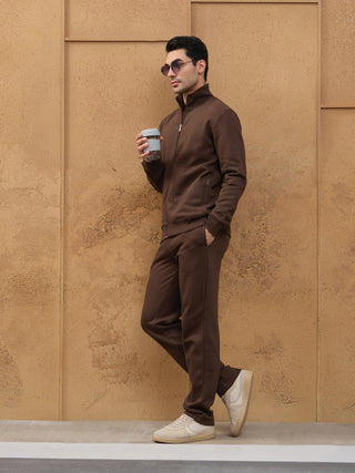 Men in a brown zippered sweatshirt and pants, standing and holding a grey cup.