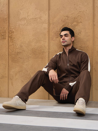 Men wearing a brown tracksuit with white stripes, sitting against a textured wall.