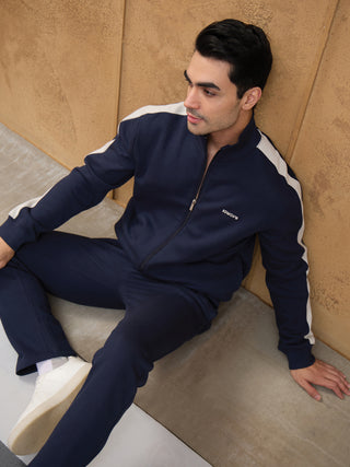 A man poses in a navy blue tracksuit with white accents, featuring a zip-up jacket and matching pants, a logo printed on the jacket is a new detail.