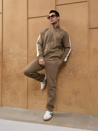 A man stands leaning against a textured wall wearing a brown tracksuit with white stripes on the sleeves and collar.