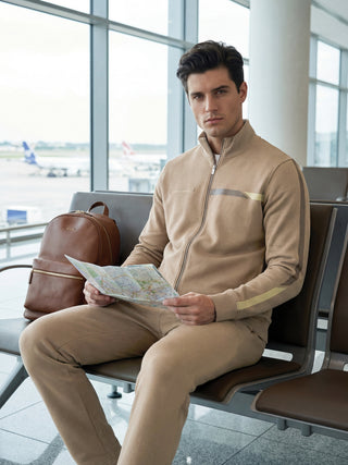 Men wearing a tan zippered tracksuit with tape detailing on the sleeves and chest is sitting in an airport and looking at a map.