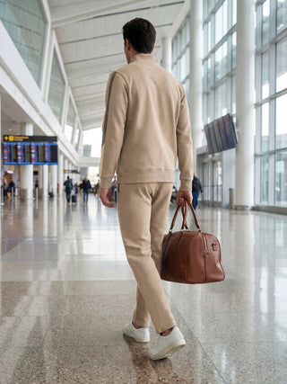 Men walking in an airport, wearing a beige tracksuit and carrying a brown duffel bag.