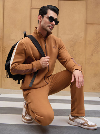 A man kneels in a brown fleece tracksuit, with white and brown accents on the sleeves and pants, and a black backpack.
