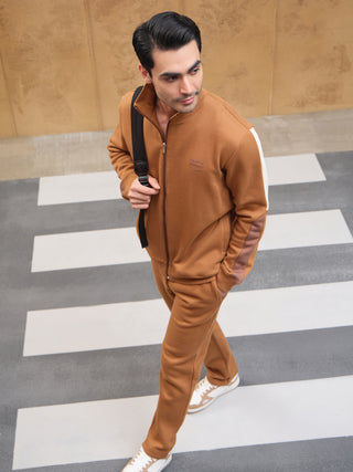 Men walking on a crosswalk wearing a brown zip-up tracksuit with white accents on the sleeves, carrying a black backpack.