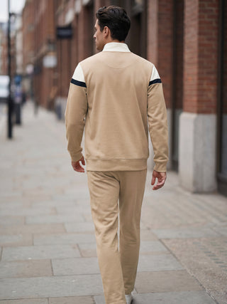 Men walking away from camera wearing a beige tracksuit with white and navy detailing.
