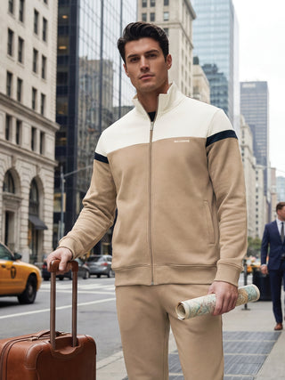 A man stands on a city street, holding a suitcase and a rolled-up map, wearing a color-blocked tracksuit in beige and cream.