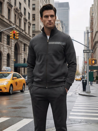 Men in a charcoal grey full-zip tracksuit with a subtle striped detail on the chest, posing with hands in pockets on a city street.