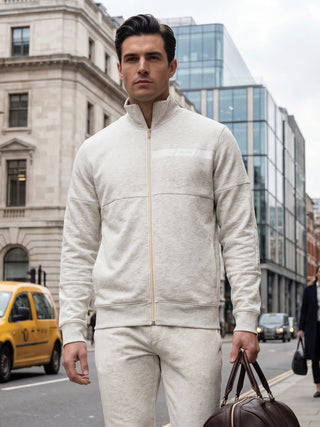 A man in a beige tracksuit with a zip-up jacket and matching pants holds a brown duffel bag.