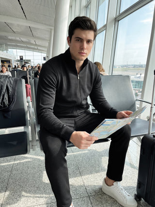 A man wearing a black waffle knit loungewear set sits in an airport lounge holding a map.