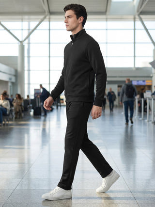 A man in a black textured zipper jacket and black textured pants, wearing white sneakers, walking in an airport.