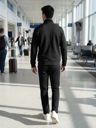 Men's black textured quarter-zip jacket and matching pants walking away from the camera in an airport.