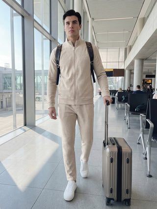 A man wearing a beige loungewear set with a zip-up jacket and matching pants, complete with a backpack and suitcase, walks through an airport.