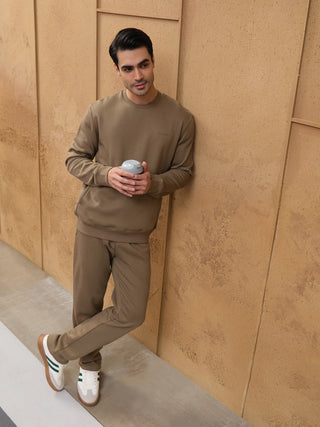 A man wearing a brown crewneck sweatshirt and matching sweatpants leans against a textured wall, holding a grey cup.