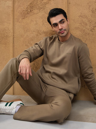 A man wearing a brown crewneck sweatshirt and matching sweatpants leans against a textured wall.