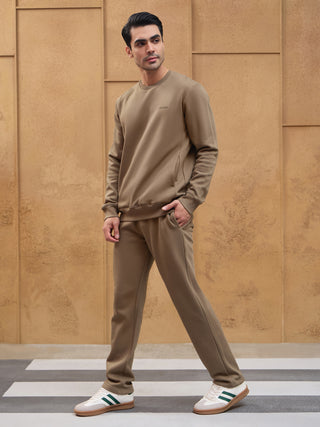 A man in a taupe sweat suit with white and green sneakers stands against a textured wall, with one hand in his pocket.