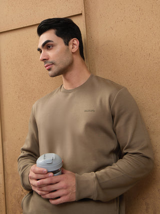 Men in a relaxed pose holding a water bottle, wearing a tan crewneck sweatshirt with a small embroidered logo.