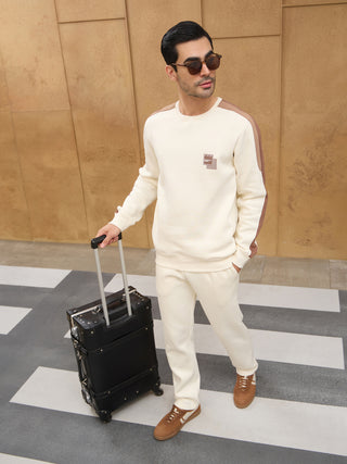 Men wearing a cream sweatshirt with brown stripe details and matching cream sweatpants, pulling a black suitcase.