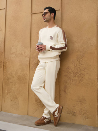 Men in a cream-colored, two-piece loungewear set with brown accents stand relaxed, holding a coffee cup.