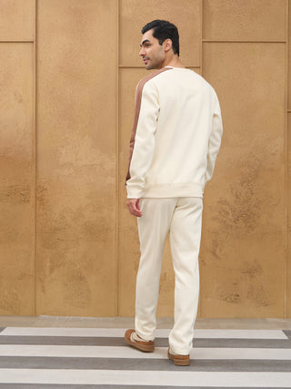 A man poses from the back wearing a cream and brown colorblock crewneck sweatshirt and cream sweatpants.