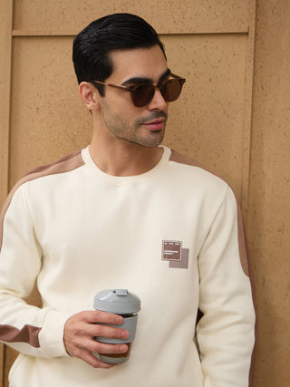 Men wearing a cream sweatshirt with brown accents and a graphic print, holding a coffee cup.