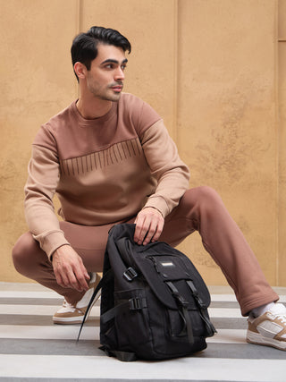 A male model squats, wearing a two-toned brown sweatshirt with a line print and matching sweatpants, with a black backpack beside him.