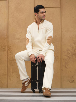 A man in a cream-colored loungewear set with brown suede elbow patches and a quarter-zip neckline sits on a black suitcase.